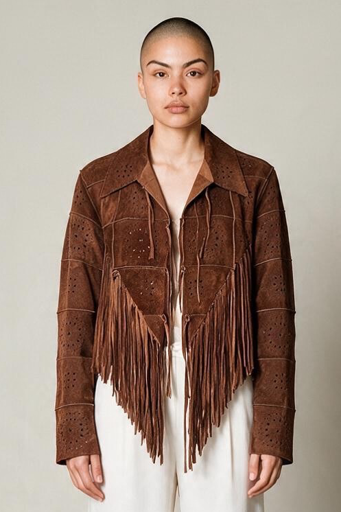 A model stands facing forward wearing a brown suede fringe jacket over a white wide-leg pants set, photographed in a minimalist studio with a warm off-white wall and gray floor.